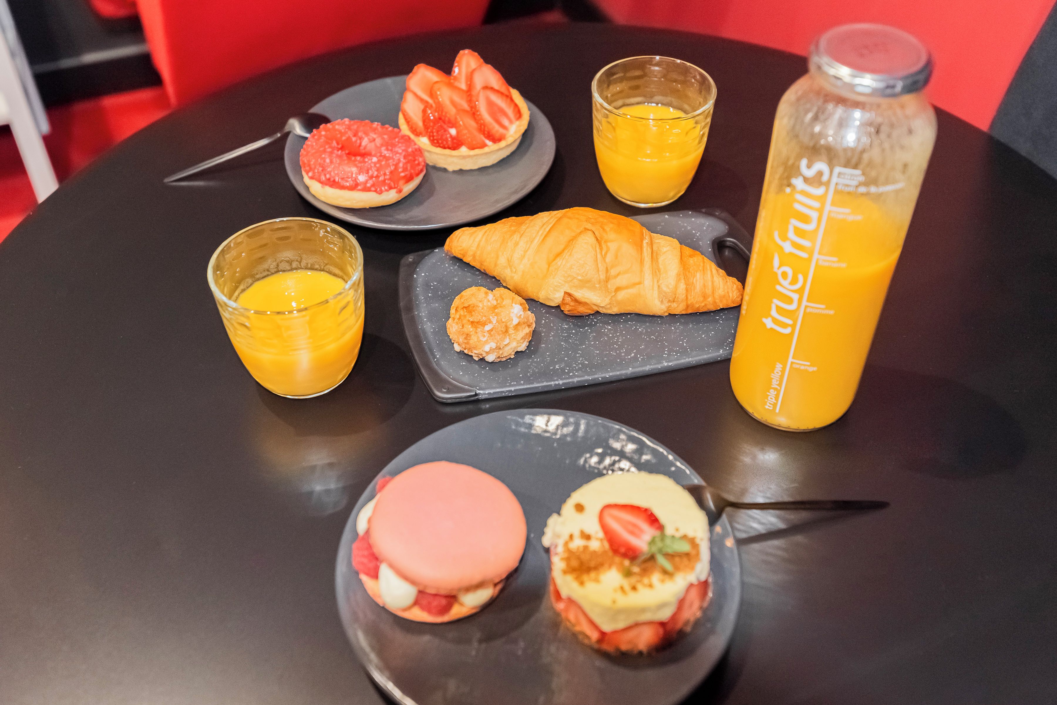 Taula amb un croissant, una tartaleta de maduixa, un got de suc d'aranja, un pot de melmelada i un postre amb una maduixa a un plat.