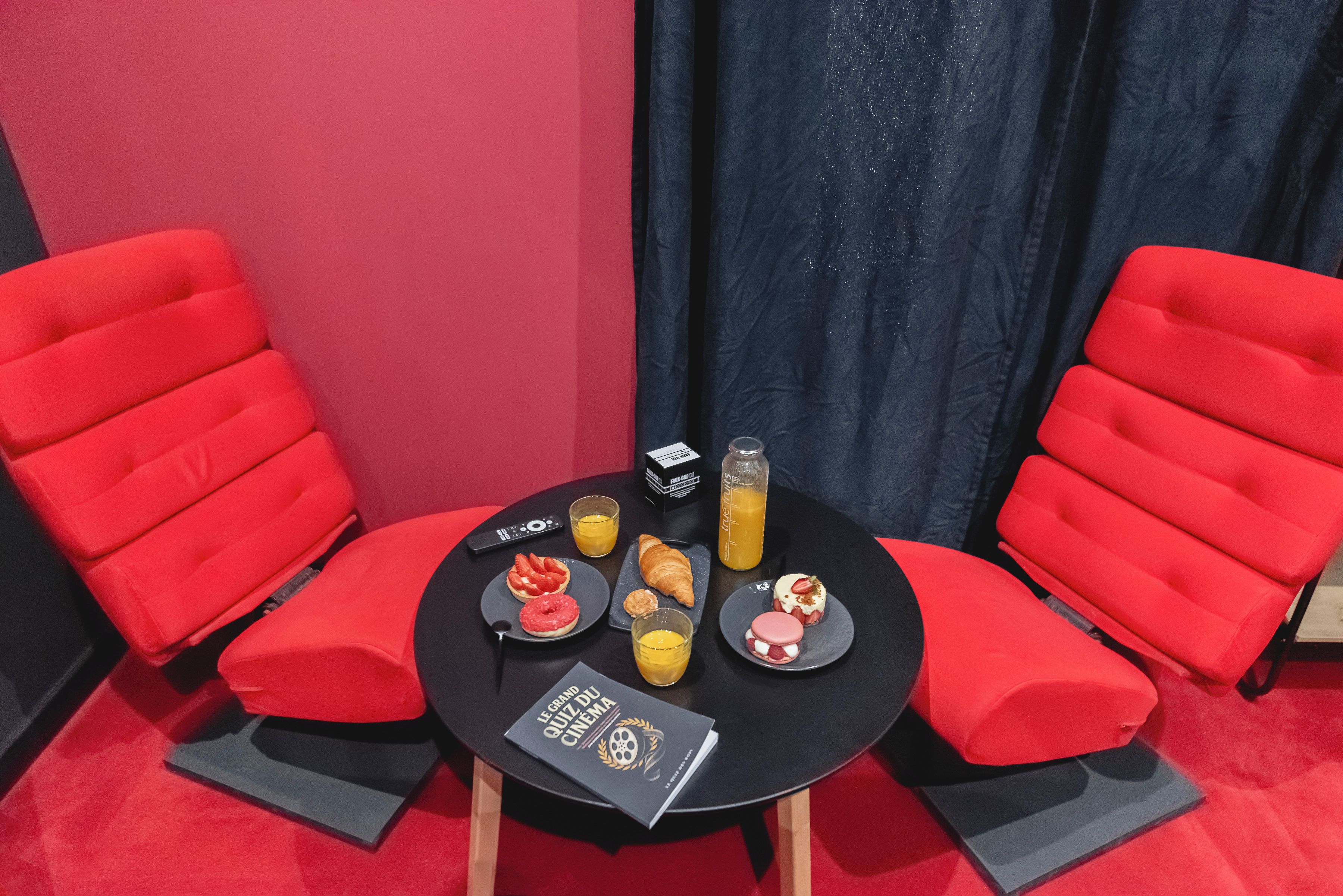 Una habitación con dos sillas rojas, una mesita con provisiones para el desayuno, un control remoto y una botella de zumo de naranja.
