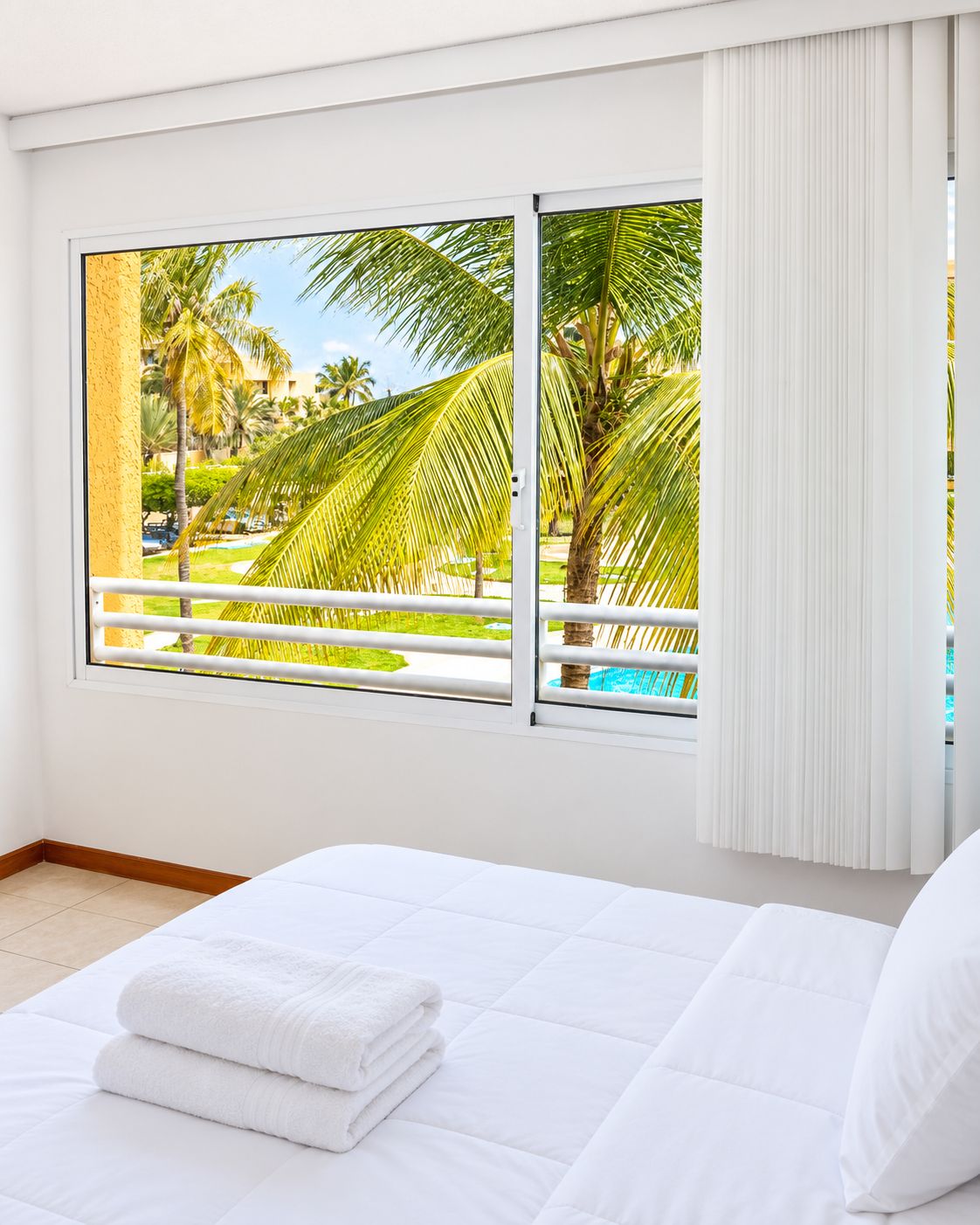 Une chambre d’hôtel avec un lit, du linge de lit blanc et, par la fenêtre, une vue sur une piscine et des palmiers.