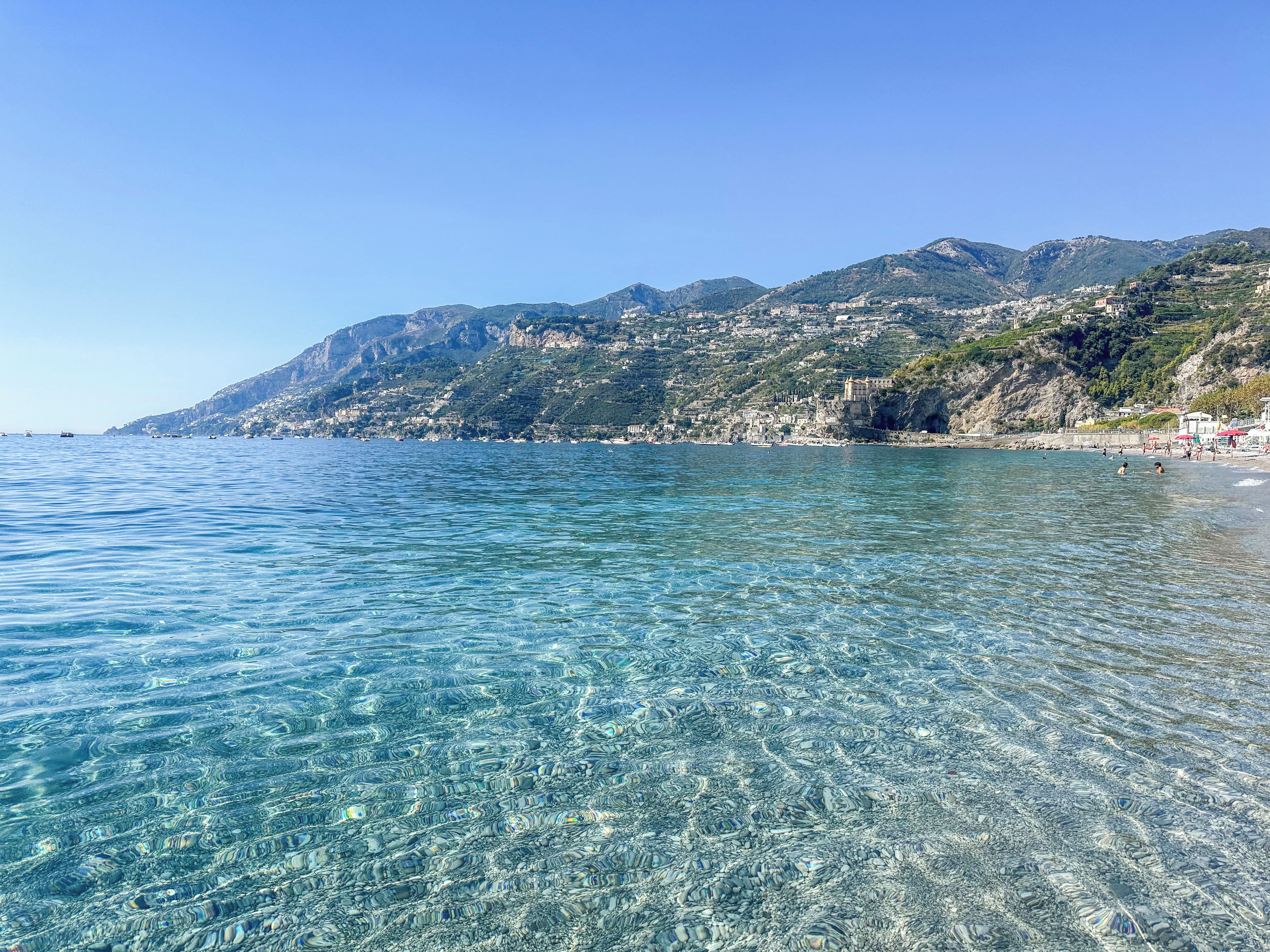A coastal view with clear water, a beach, and a hillside with buildings.