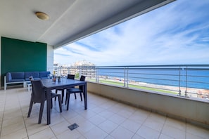 A balcony with a dining table, chairs, and a sofa. The balcony overlooks a body of water and a distant building. - Oceanview Luxury, Fort Cambridge W/pool (Sliema)