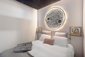 A modern bedroom with a circular moon-like wall art, a bed with white linens, and two wall-mounted light fixtures.