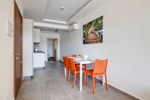 Un coin repas avec une table blanche et des chaises orange, une cuisine en arrière-plan et un tableau accroché au mur.