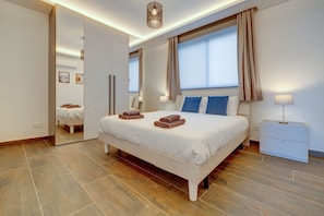 A modern bedroom with a large bed, a bedside table, a mirror, and a window with curtains. - Idyllic Apartment With Valletta and Harbour Views (Gzira)
