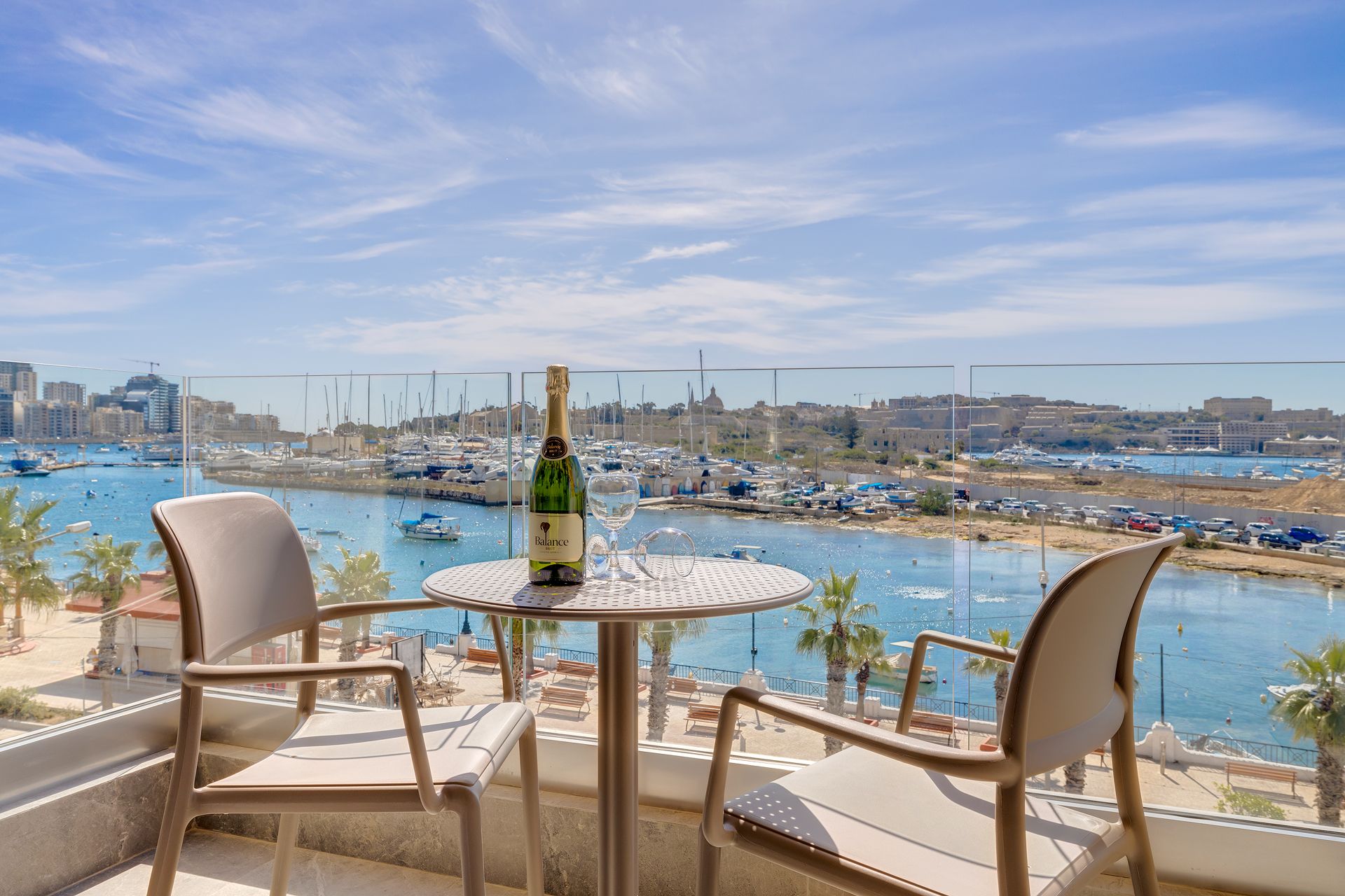 A balcony with a table and chairs overlooking a marina with boats and a cityscape.