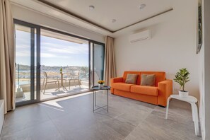 A modern living room with a large sliding glass door leading to a balcony, an orange sofa, a small table, and a potted plant. - Idyllic Apartment With Valletta and Harbour Views (Gzira)