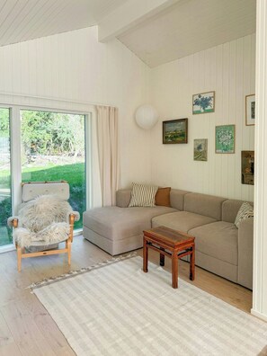 A cozy living room with a sofa, a chair, a small table, and a view of greenery through a large window. - Newly built dream summer house on Sjællands Odde (Sjællands Odde)