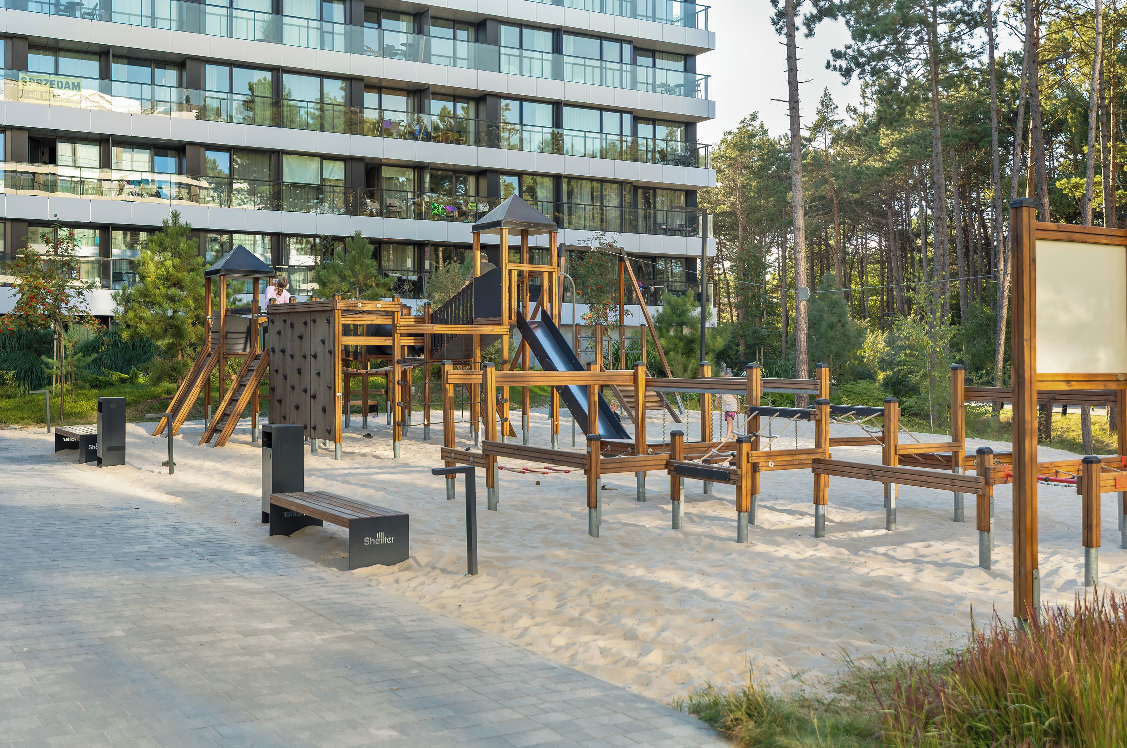 A playground with various play structures, including slides and climbing areas, set against a backdrop of a modern building and a forested area.