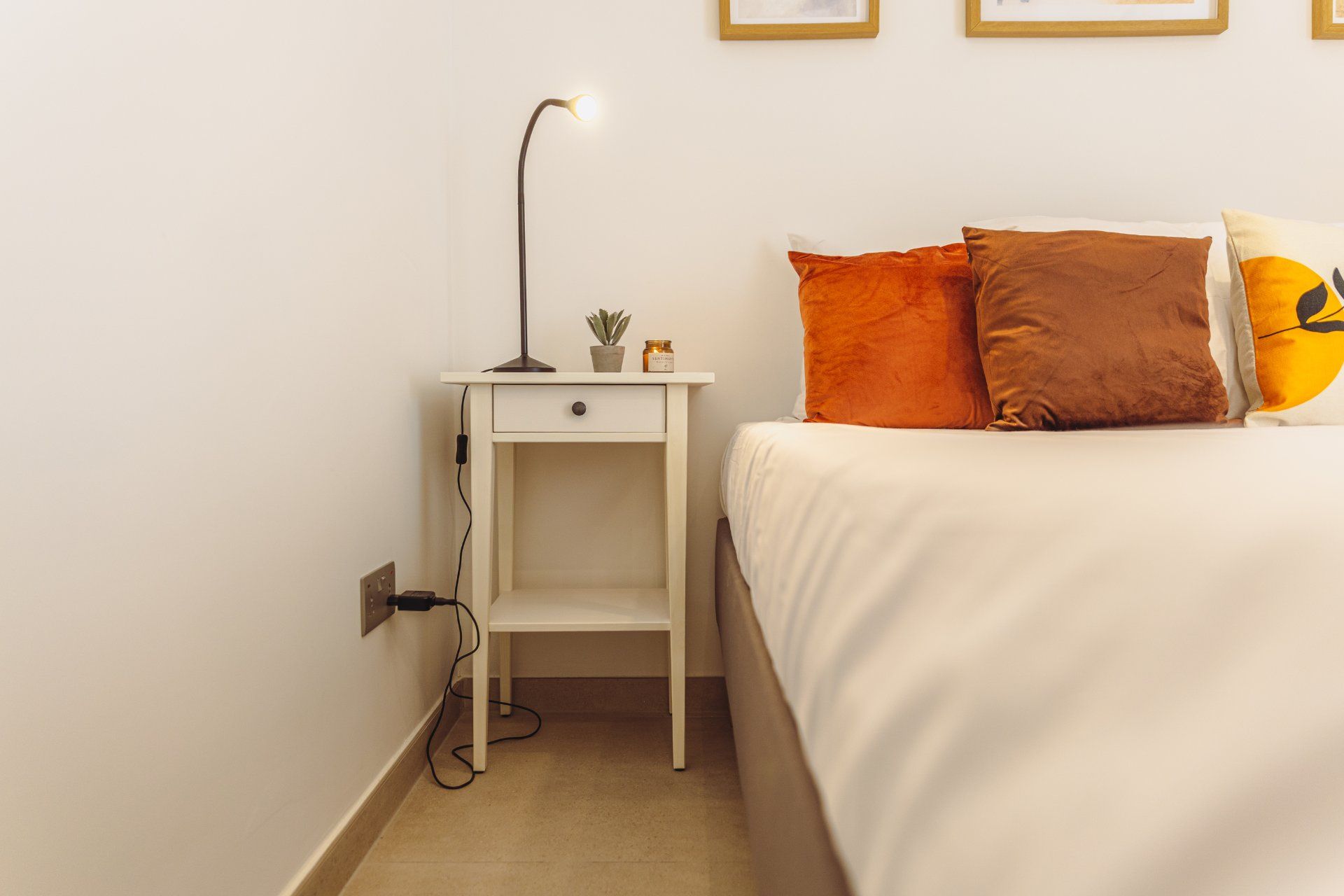 A neatly arranged bedroom with a white bed, orange and brown pillows, a bedside lamp, and a small plant on a nightstand.