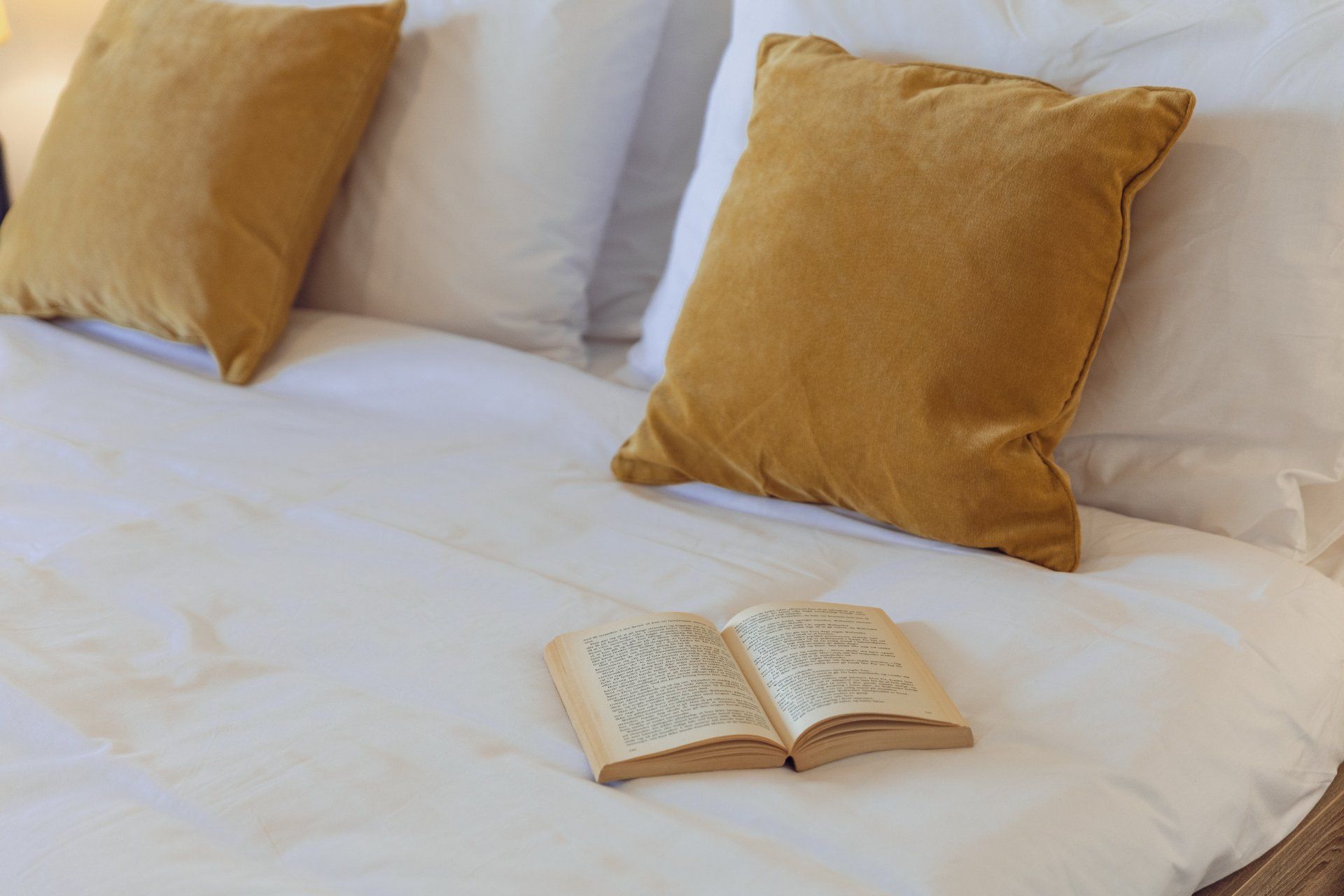 A bed with white bedding and two mustard-colored pillows, an open book on the bed.