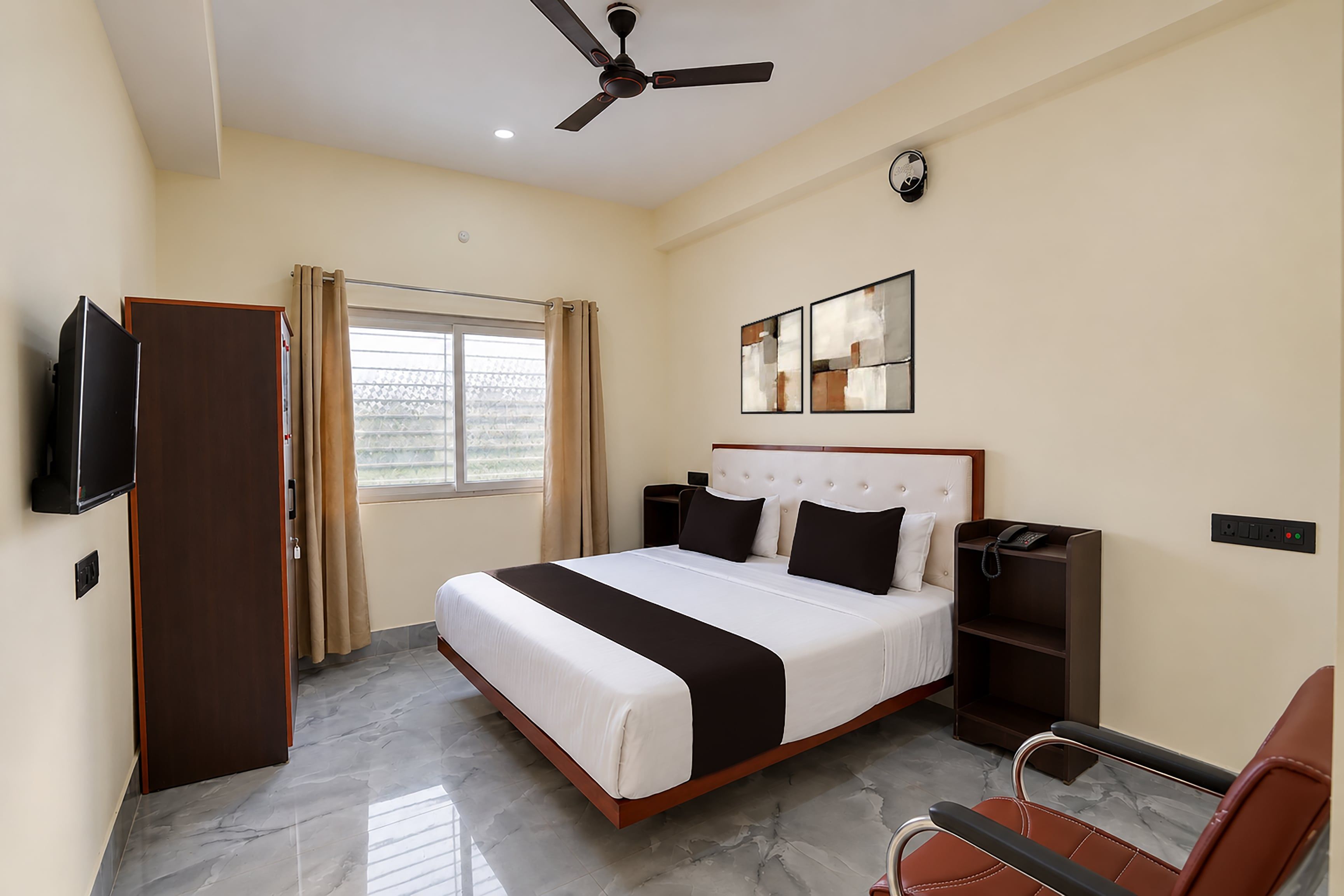 A hotel room with a bed, a TV, a ceiling fan, a window with curtains, and a bedside table with a phone.