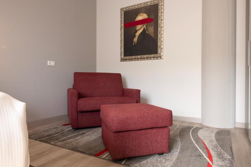 Quarto com poltrona e banqueta vermelhas, um retrato emoldurado na parede e um tapete estampado.