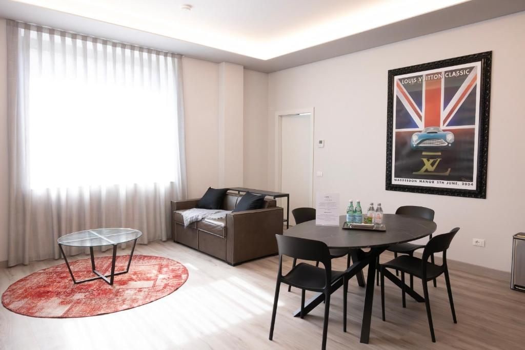 Un salon moderne avec une table à plateau en verre, un canapé et une table à manger avec des chaises. Il y a une affiche au mur et une fenêtre avec des rideaux.