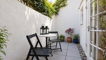 A small outdoor patio with a round table and two chairs, surrounded by potted plants and a white wall.