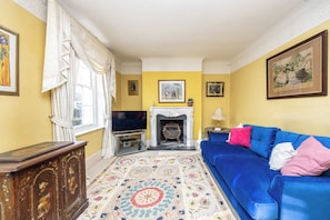 A living room with a fireplace, a blue sofa, a television, and a patterned rug. - Seaside Historic Townhouse, 1 Min to Beach, (Worthing)