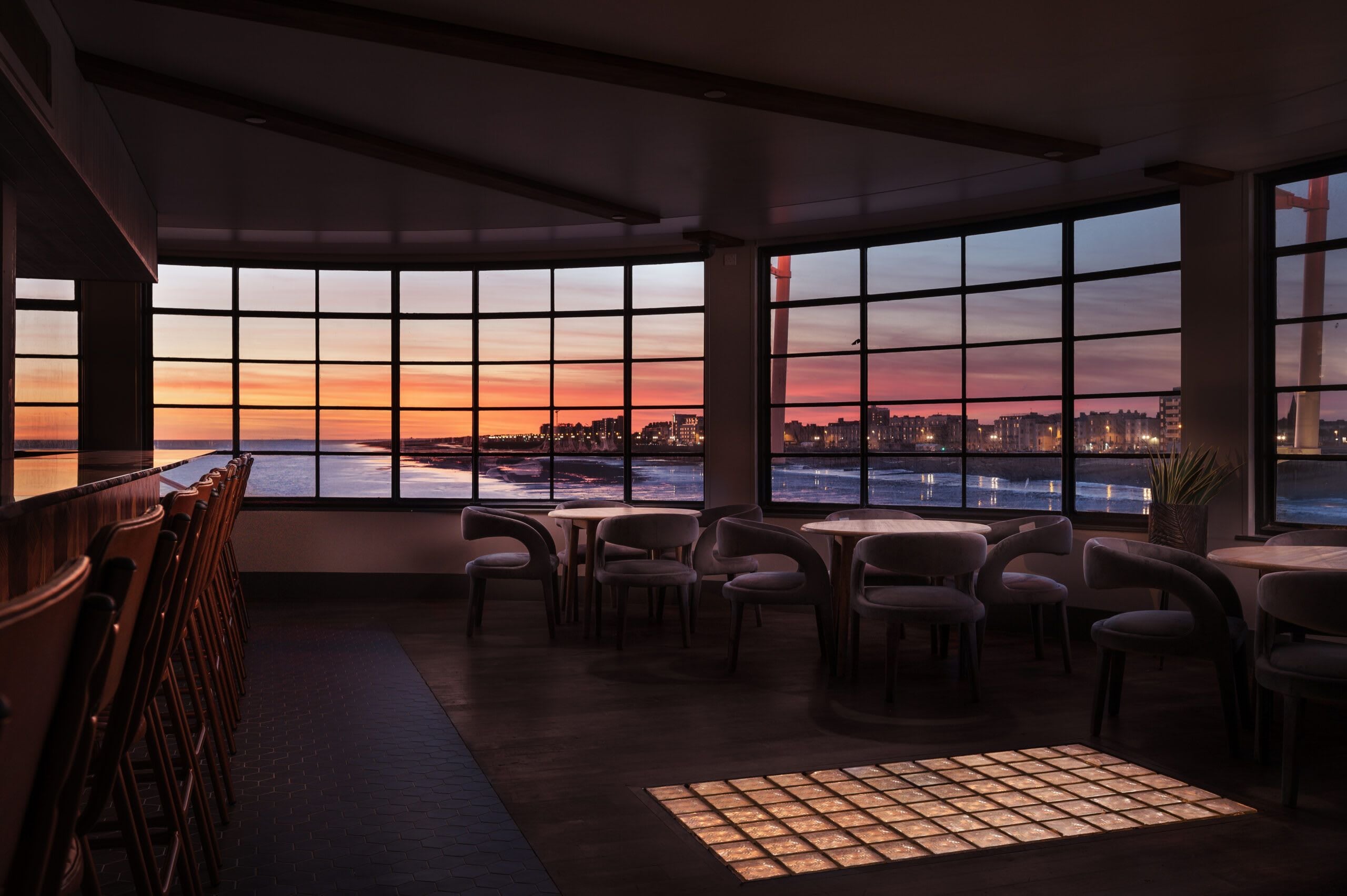 A restaurant with large windows offering a city view at sunset.