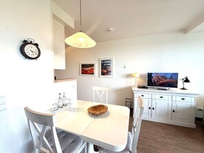 A modern kitchen with white cabinetry, a dining table with chairs, a clock, a pendant light, and a television on a cabinet. - Halligblick Apt. 11 (Wyk auf Foehr)