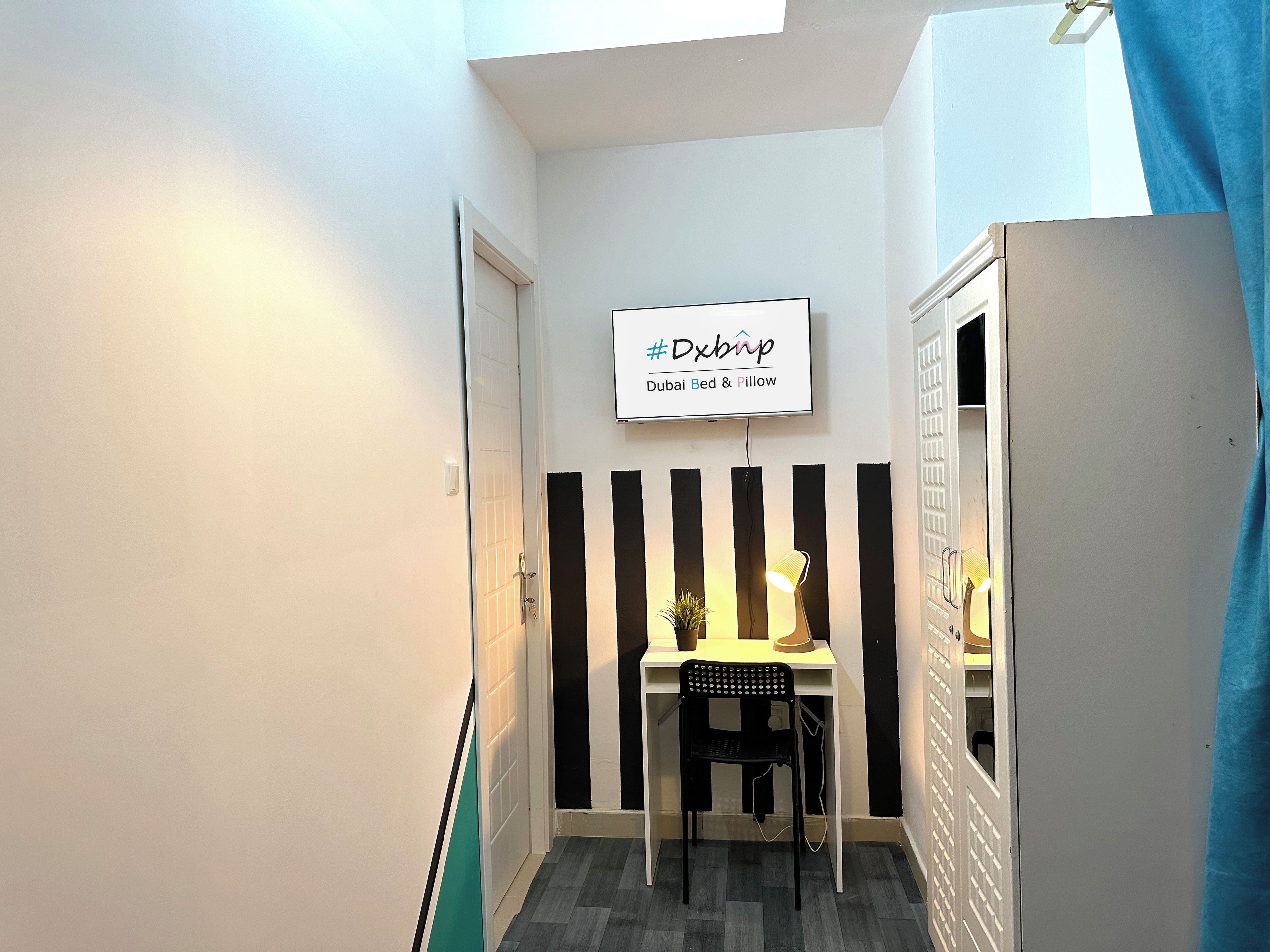 Een smalle gang met een bureau en stoel, een bordje met de tekst 'Dubai Bed & Pillow' en een deur aan de linkerkant.