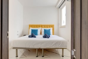 Une chambre d’hôtel avec un lit, du linge de lit blanc, une tête de lit jaune et deux oreillers bleus.