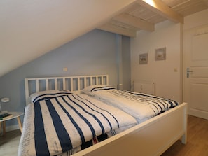 Ein Schlafzimmer mit schrägem Dach, einem weißen Bett, gestreifter Bettwäsche, einem kleinen Nachttisch und einer Heizung.
