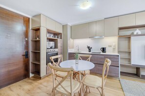 A compact kitchen with a dining area, wooden cabinets, a built-in oven, and a round table with chairs. - Ñuñoa Botanic 2D - Estacionamiento y AC (Ñuñoa)