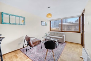 Un salon moderne comprenant un canapé, une table basse, un fauteuil, un tapis à motifs, un luminaire suspendu et une grande fenêtre donnant sur des immeubles.