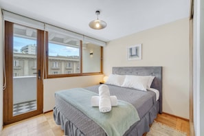 A bedroom with a bed, a bedside lamp, a window with a view of buildings, and a door with a telephone. - Acogedor departamento bohemio en Providencia (Providencia)