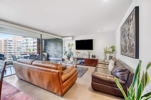 A modern living room with leather sofas, a flat-screen TV, and a large window overlooking a cityscape. - Depto. 3D Prime Spot: Parque Arauco + Metro (Las Condes)
