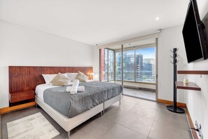 Een moderne hotelkamer met een groot bed, een houten hoofdbord, stadszicht door raam-tot-plafond ramen, een flatscreen televisie aan de muur bevestigd, en een klein houten plankje.
