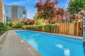 A rectangular swimming pool with a paved walkway on one side, surrounded by a brick wall and a wooden fence. - Depto. 3D Prime Spot: Parque Arauco + Metro (Las Condes)