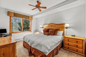 Quarto com cama, mesas de cabeceira, escrivaninha, ventoinha no teto e janela com vista para árvores.