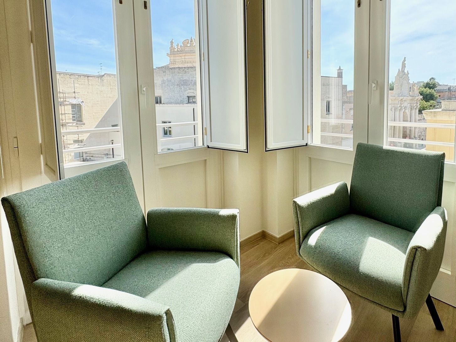A room with two green armchairs and a round table, large windows with a view of buildings and a clear sky.