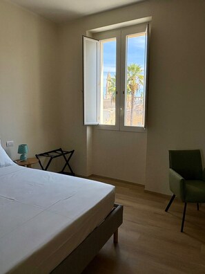 Een slaapkamer met een bed, een stoel, een lamp en een raam met uitzicht op palmbomen en gebouwen.