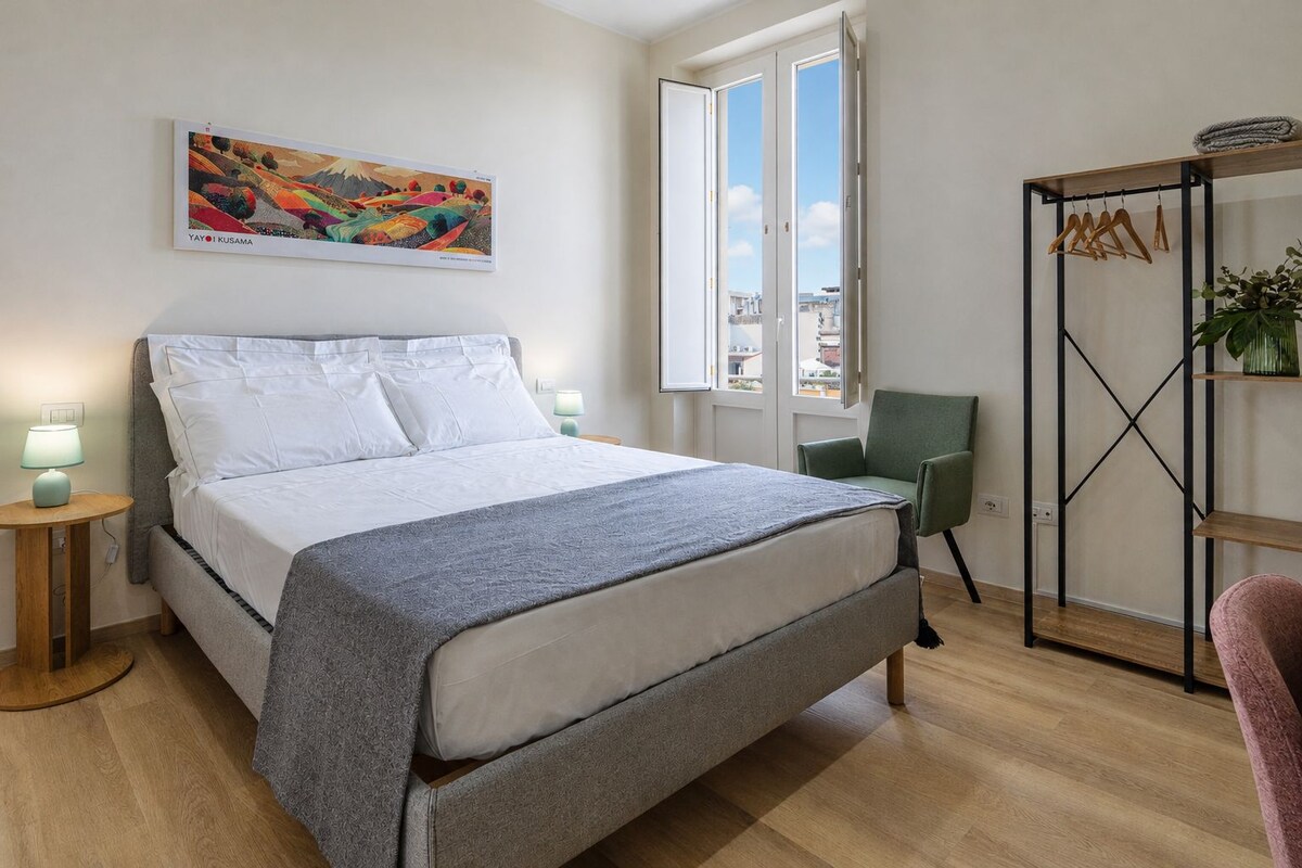 A bedroom with a bed, a chair, a wardrobe, a painting, and a window.
