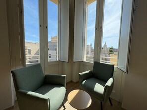 Een kamer met twee fauteuils en een kleine ronde tafel, grote ramen met uitzicht op gebouwen.