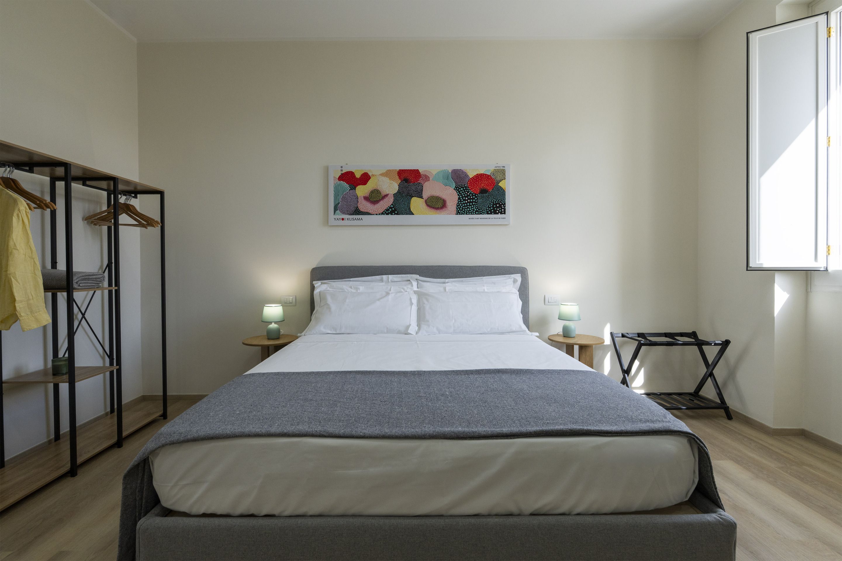 A hotel room with a bed, a wardrobe, a bedside table with a lamp, a chair, and a wall art piece.