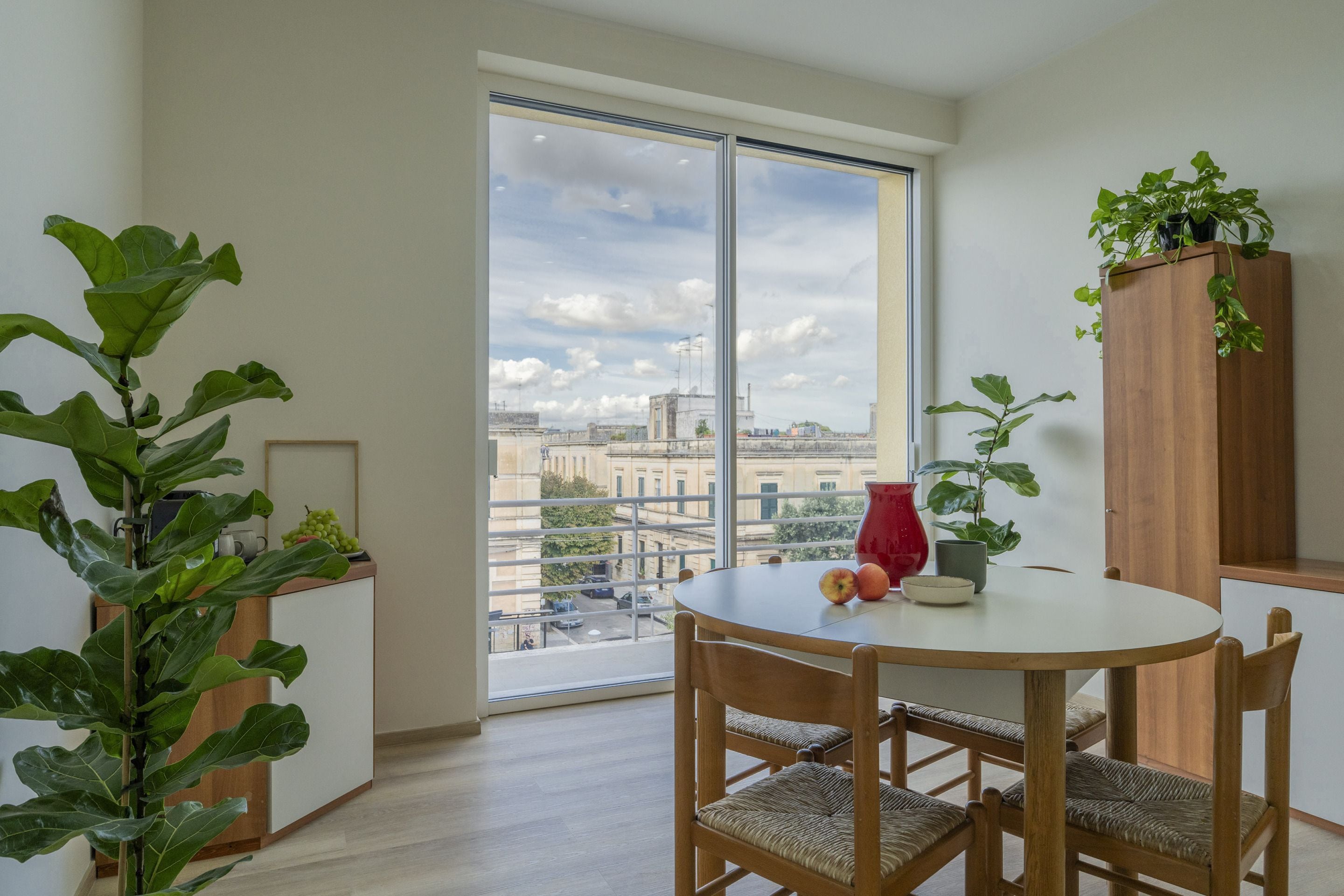 A modern apartment with a large window, a dining table with chairs, and a wooden cabinet with plants.