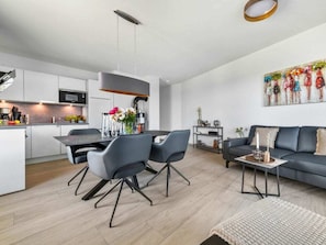 A modern kitchen with white cabinets, a dining area with grey chairs, and a living room with a sofa and a painting on the wall. - Apartment in Olpenitz With Swimming Pool (Kappeln)