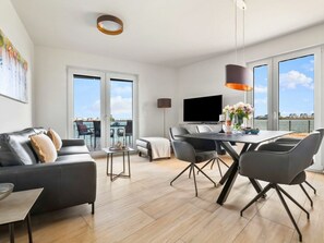 A modern living room with a sofa, a dining table with chairs, a TV, and a view of the sea through large windows. - Apartment in Olpenitz With Swimming Pool (Kappeln)