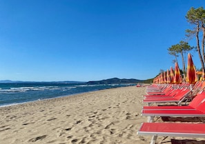 A beach with rows of red lounge chairs and orange umbrellas. - Natural Stone House with a Pool and Ocean View in the Maremma (Prata)