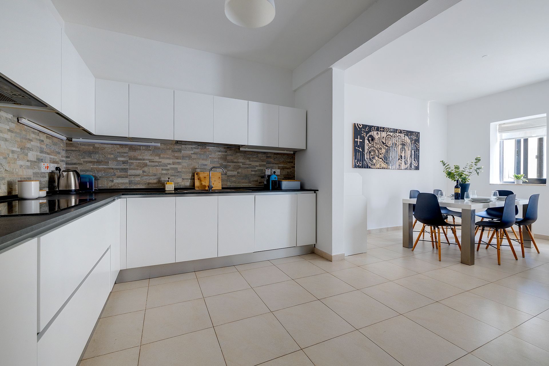 Cozinha moderna com armários brancos, bancada preta e chão revestido em azulejos. Há uma área de jantar com mesa e cadeiras.