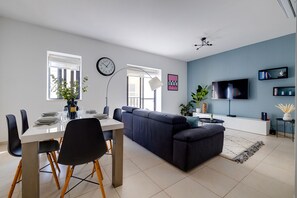 A modern living room with a grey sofa, a dining table with chairs, a TV, and a clock on the wall. - Seaside Bliss - 3BR Holiday Apartment, Sliema (Sliema)
