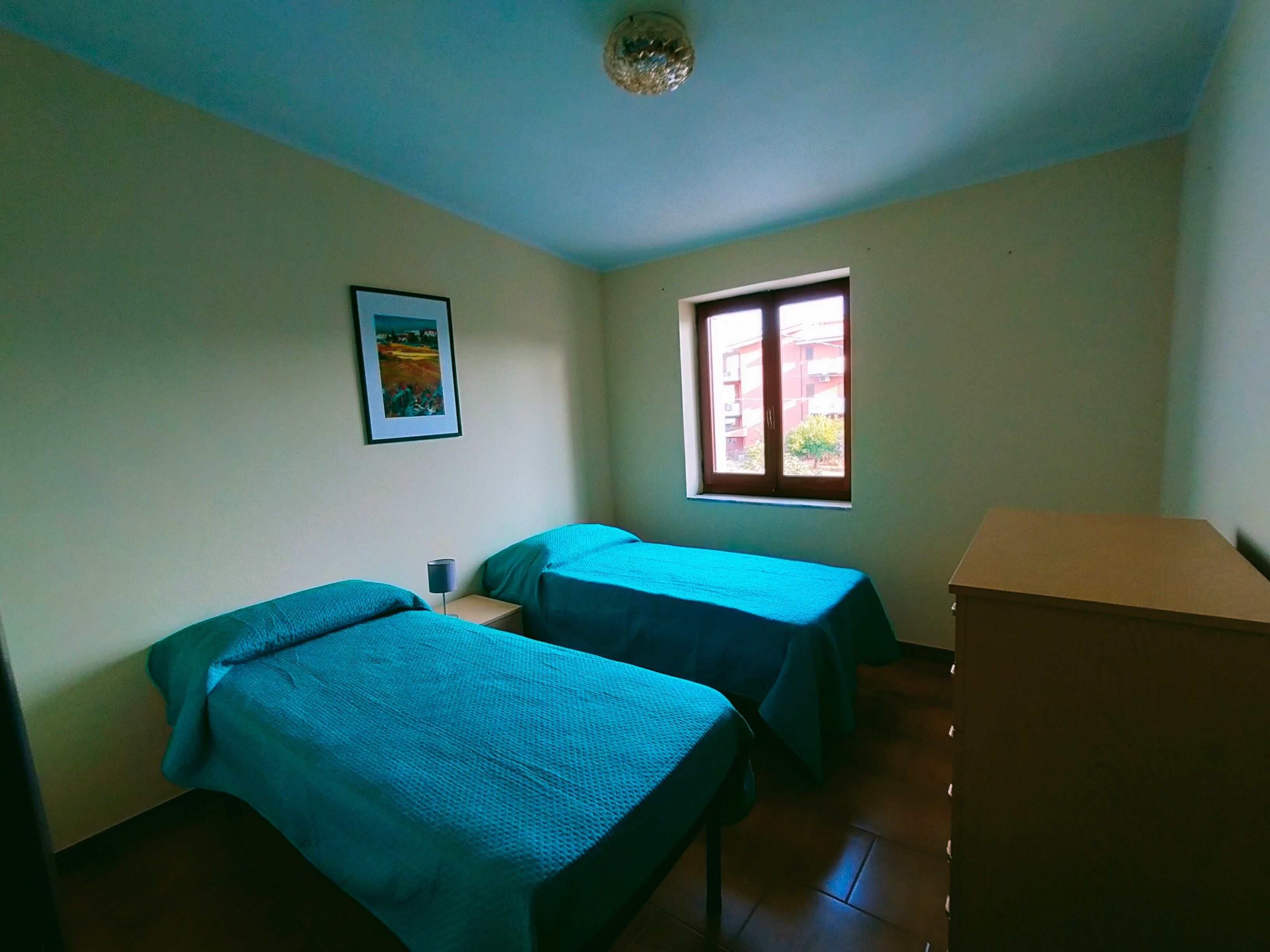 Ein Zimmer mit zwei Betten, einem Fenster, einem Bild an der Wand und einem Holzschrank.