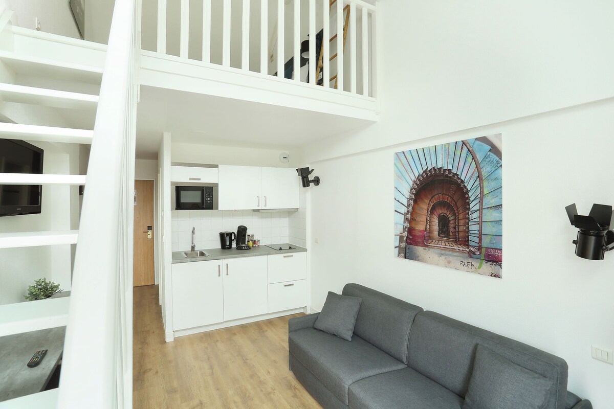 Een modern appartement met een keuken, een grijze bank en een trap naar een hoger niveau.