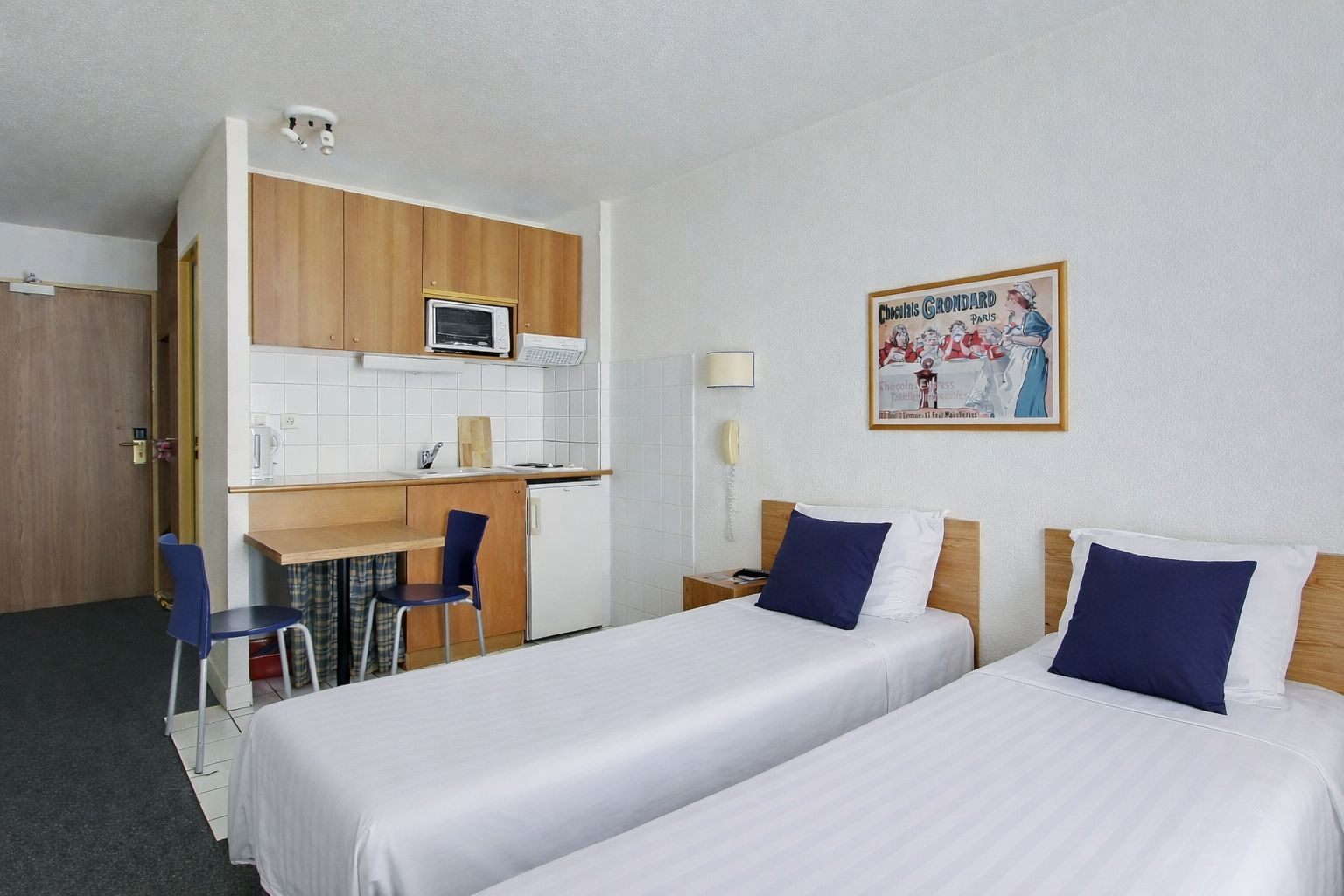A hotel room with two beds, a kitchenette, a desk, and a chair.
