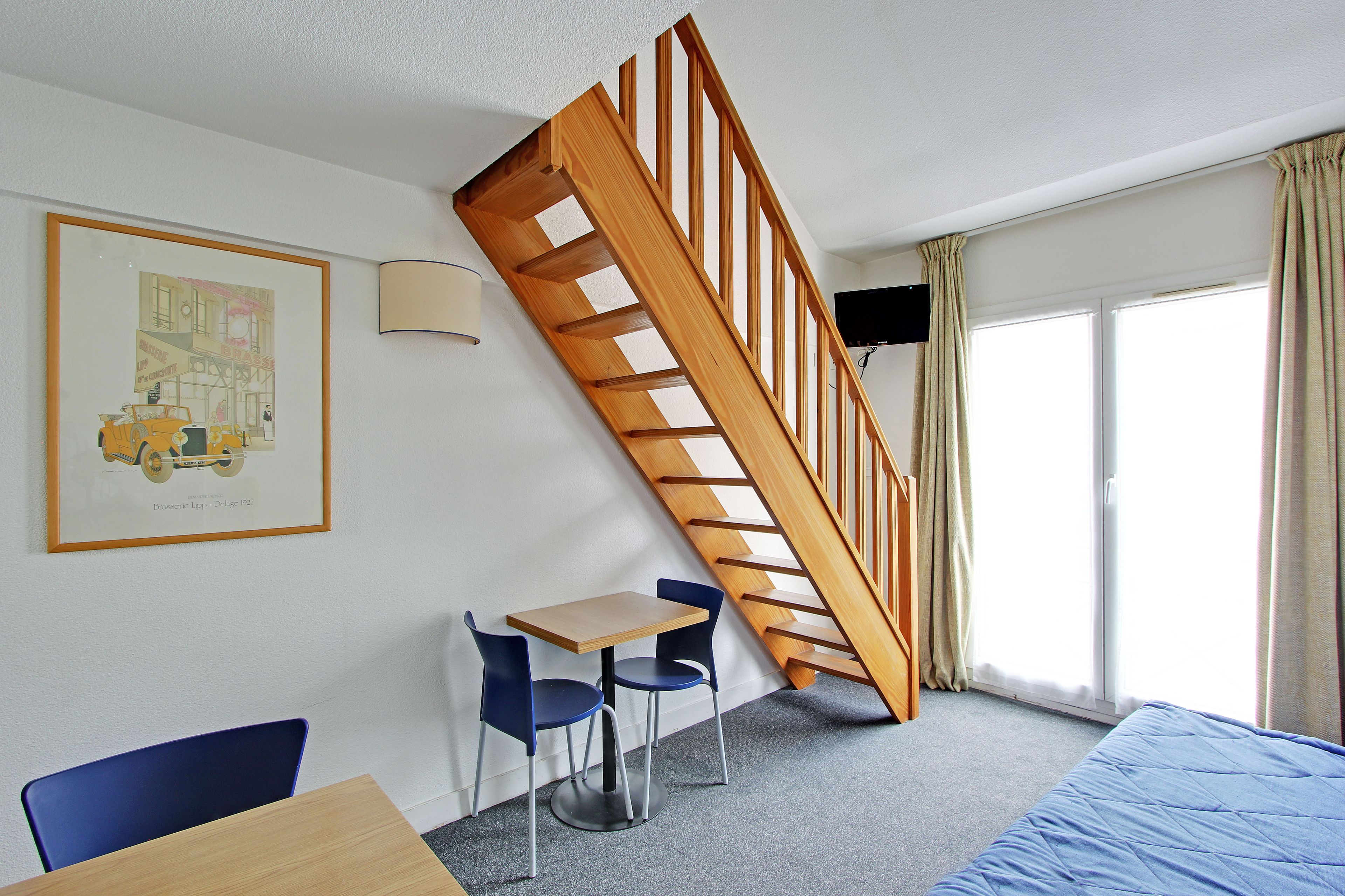 Une pièce avec un escalier en bois, un bureau, deux chaises, un lit, une fenêtre avec des rideaux et un téléviseur fixé au mur.
