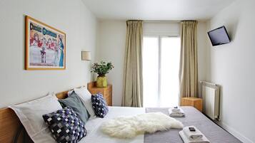 Un dormitorio con cama, almohadas, un tapete blanco y esponjoso, una mesita de noche con una planta, un televisor montado en la pared y una ventana con cortinas.