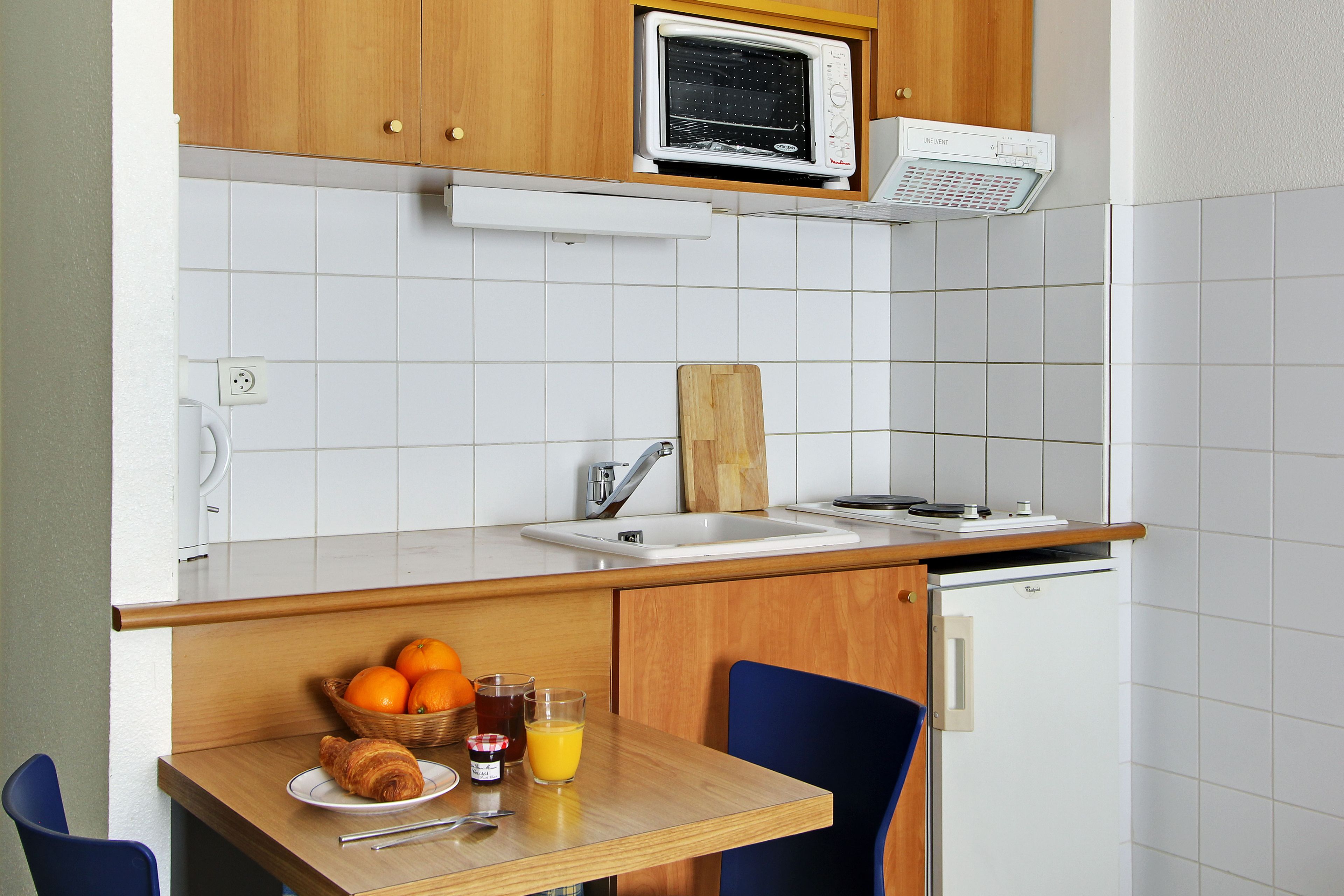 A compact kitchen with a microwave, toaster, and sink. There's a small refrigerator, a wooden table with breakfast items, and a blue chair.