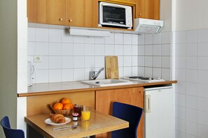 A compact kitchen with a microwave, toaster, and sink. There's a small refrigerator, a wooden table with breakfast items, and a blue chair. - YEF HOTEL LES LILAS (Les Lilas)