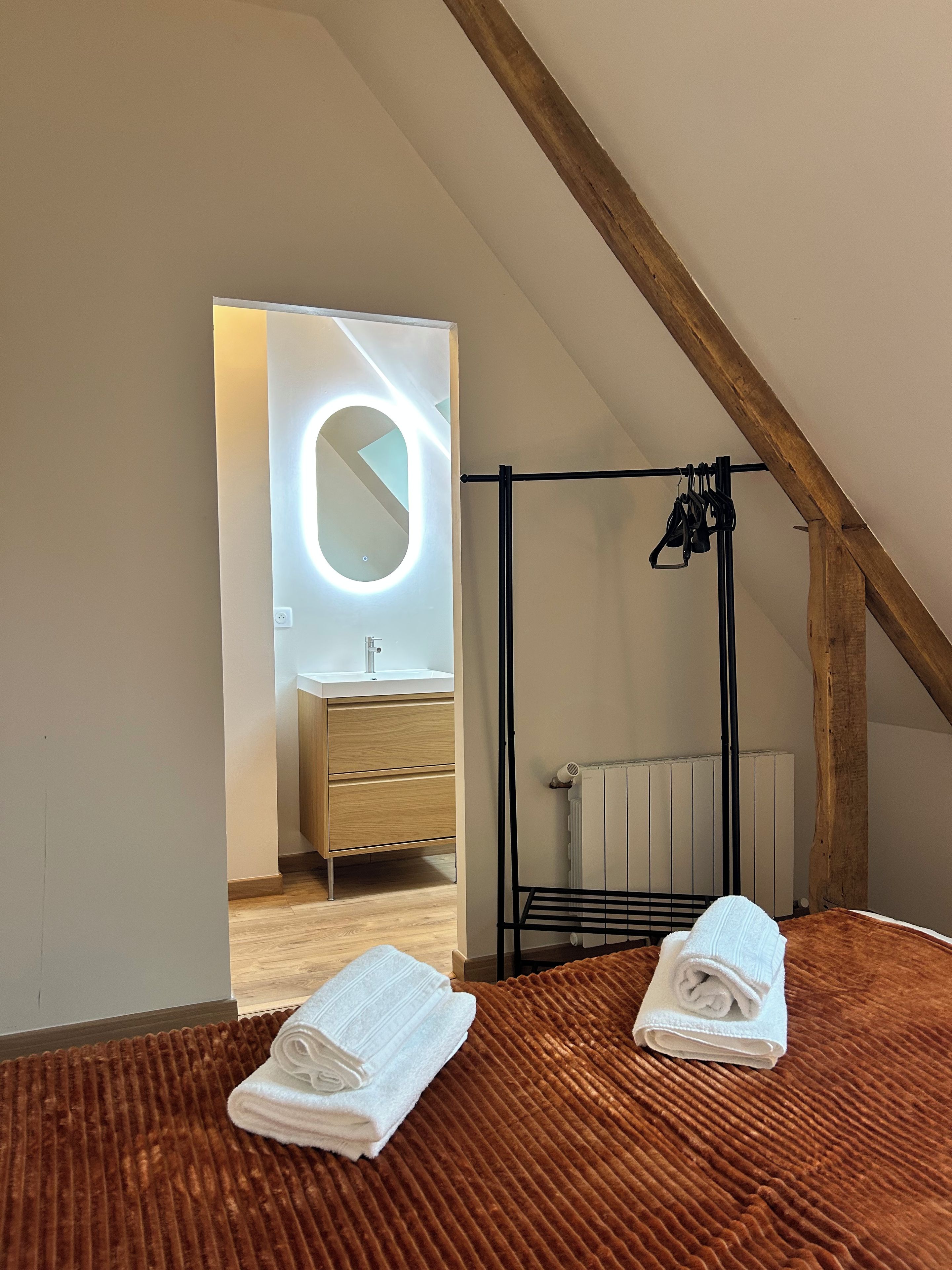 A bedroom with a wooden ceiling, a bed with a brown textured bedspread, a black metal rack, and a bathroom visible through an open door.